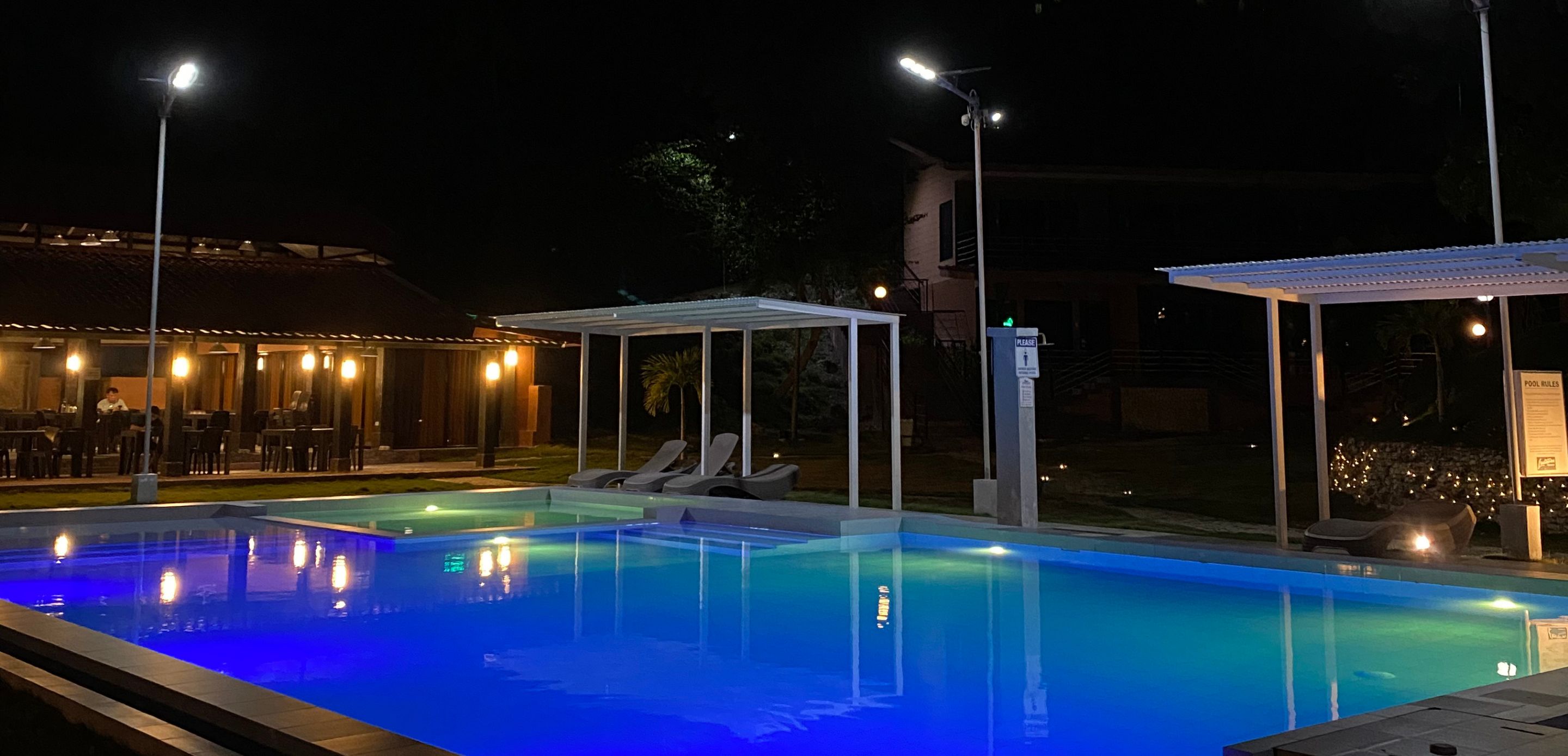 Una piscina iluminada con luces azules, sillas de descanso y un área de estar techada.