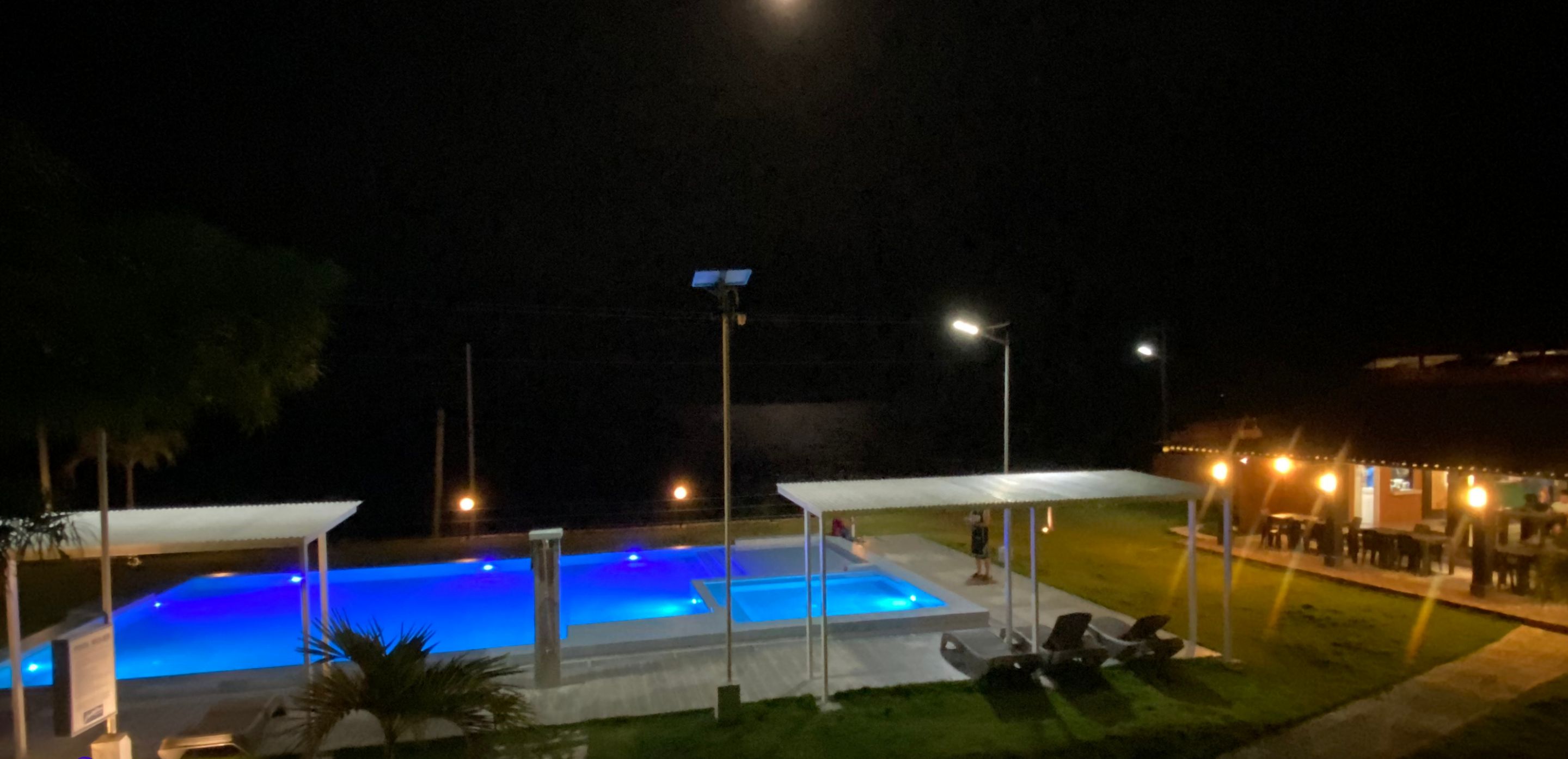 A nighttime view of a resort with a swimming pool, covered seating areas, and a well-lit outdoor dining area.