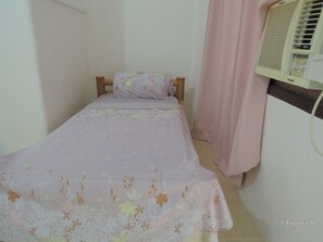 Una piccola camera con un letto singolo, copripiumino floreale, una testiera in legno, una tenda rosa e un condizionatore d'aria a parete.