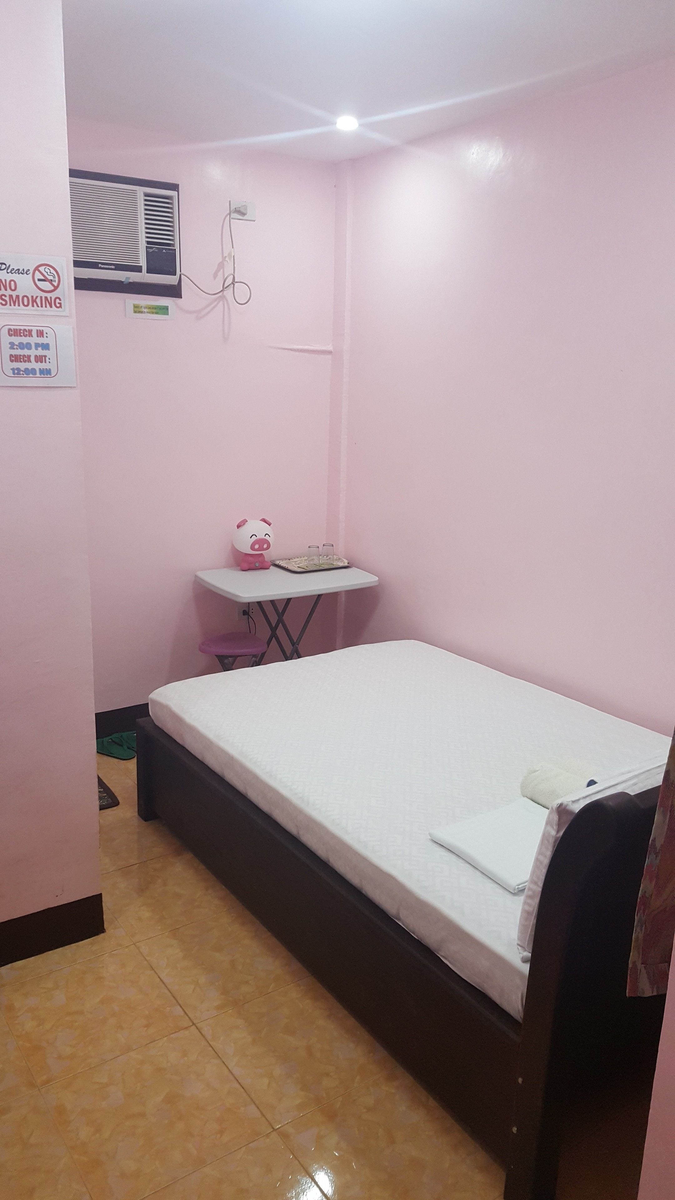 A small, pink-walled room with a single bed, a small table with a pig figurine, a heater, and a no-smoking sign.