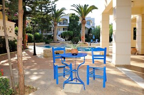 Área da piscina com mesa e cadeiras azuis, palmeiras e um edifício ao fundo.