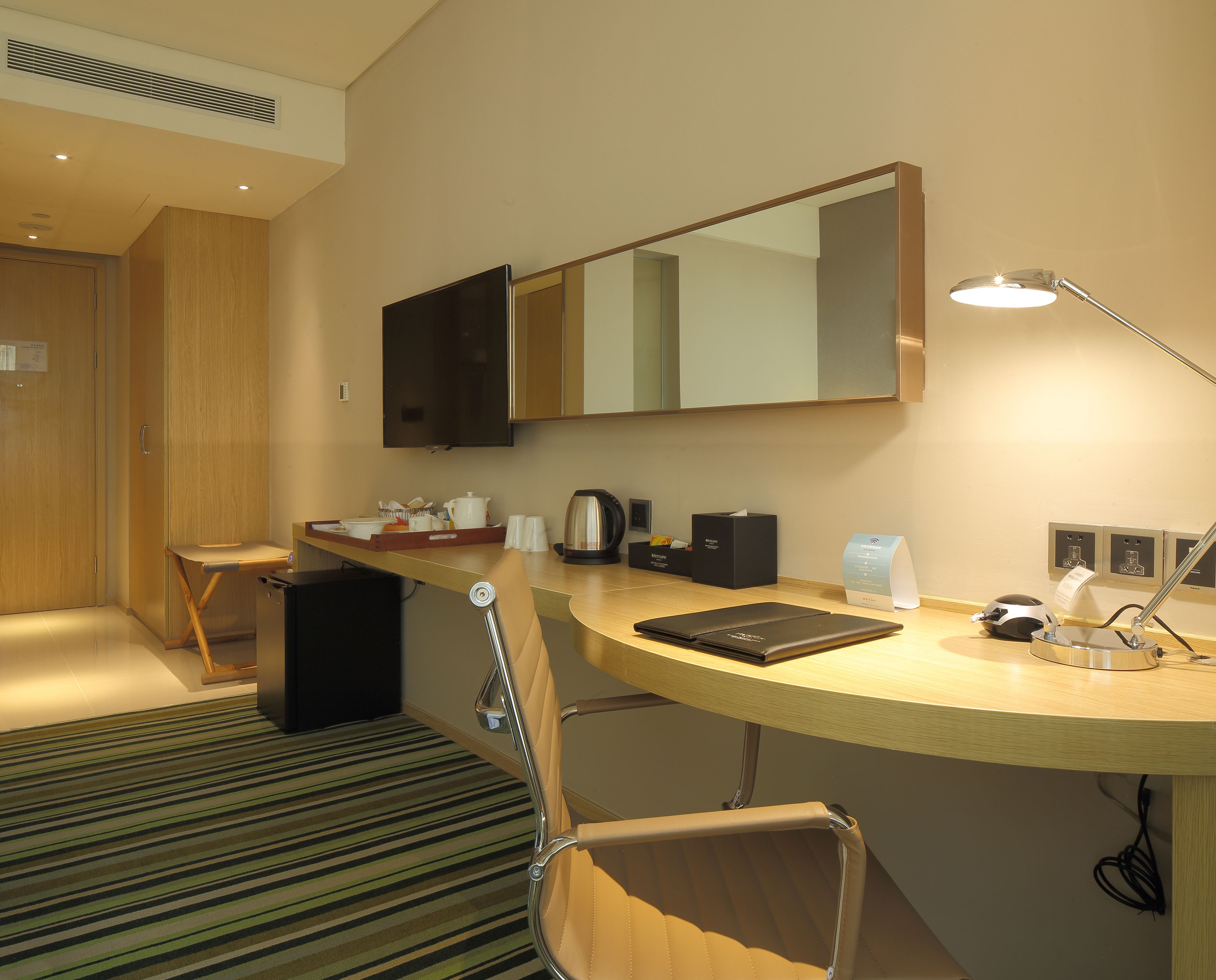 A hotel room with a wooden desk, a chair, a flat-screen TV, a mirror, a lamp, and a coffee maker.