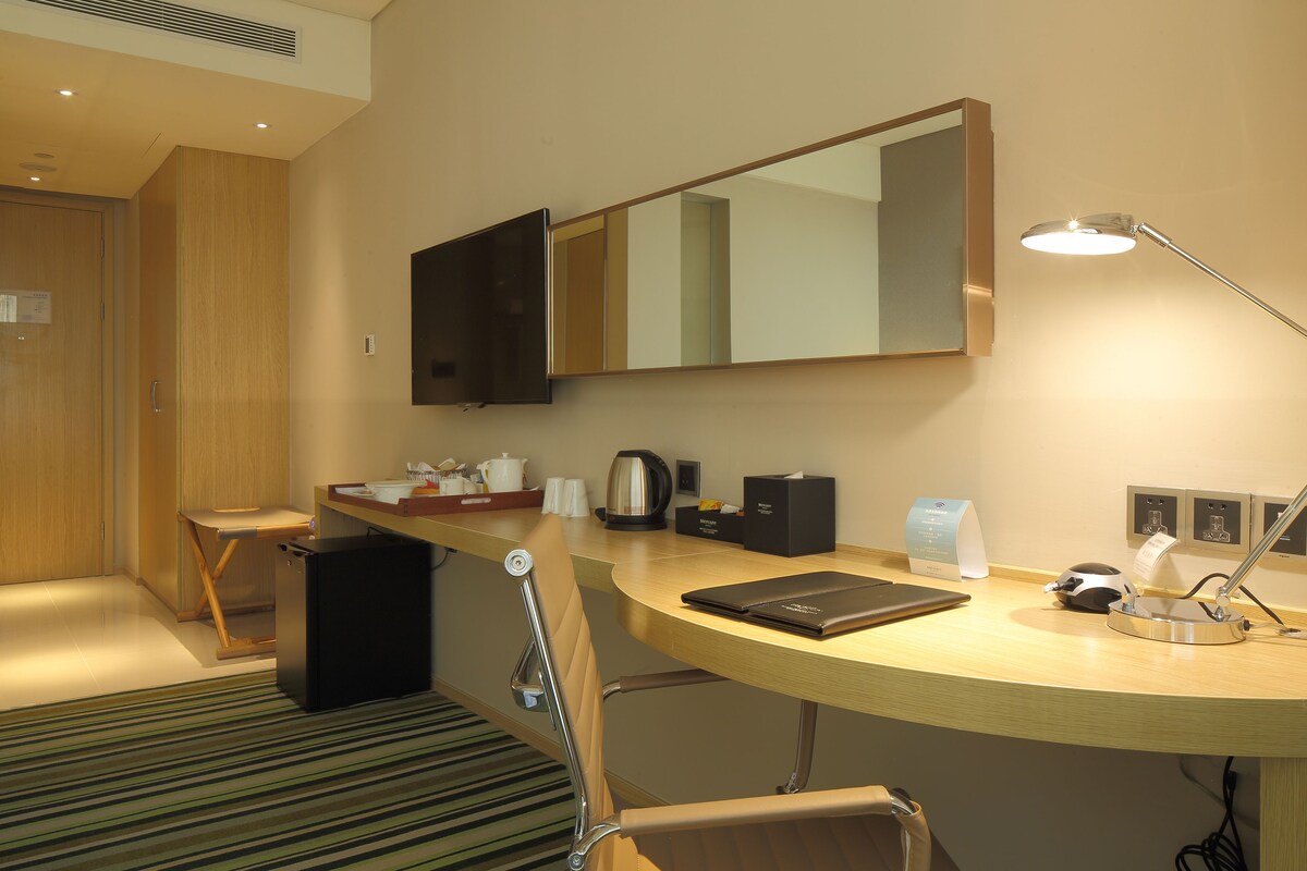Habitación de hotel con un escritorio de madera, una silla, un televisor de pantalla plana, un espejo, una lámpara y una cafetera.