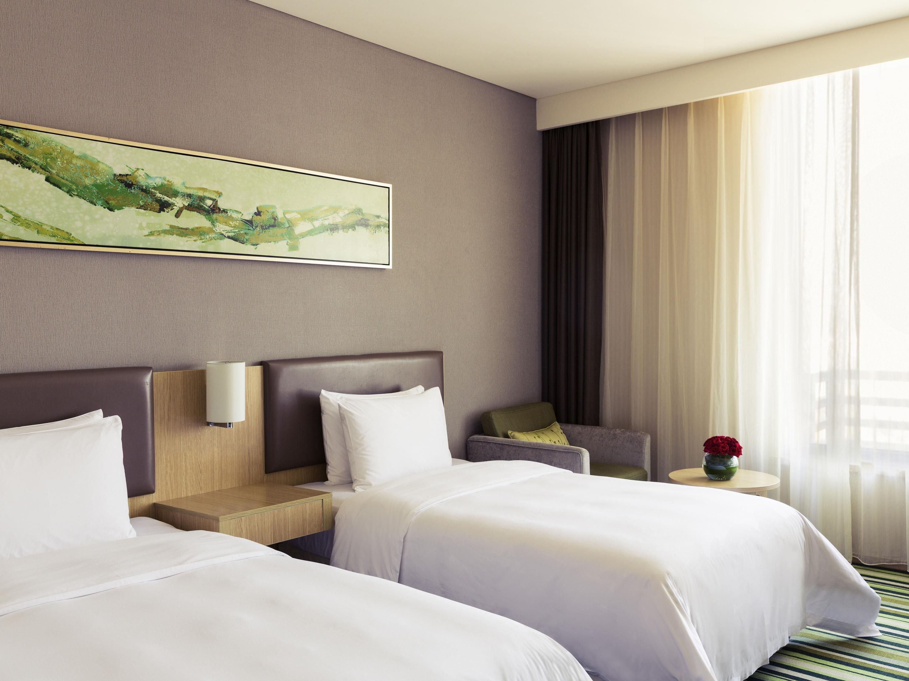 Habitación de hotel con dos camas, una mesita de noche, lámpara, silla, cuadro y ventana con cortinas.