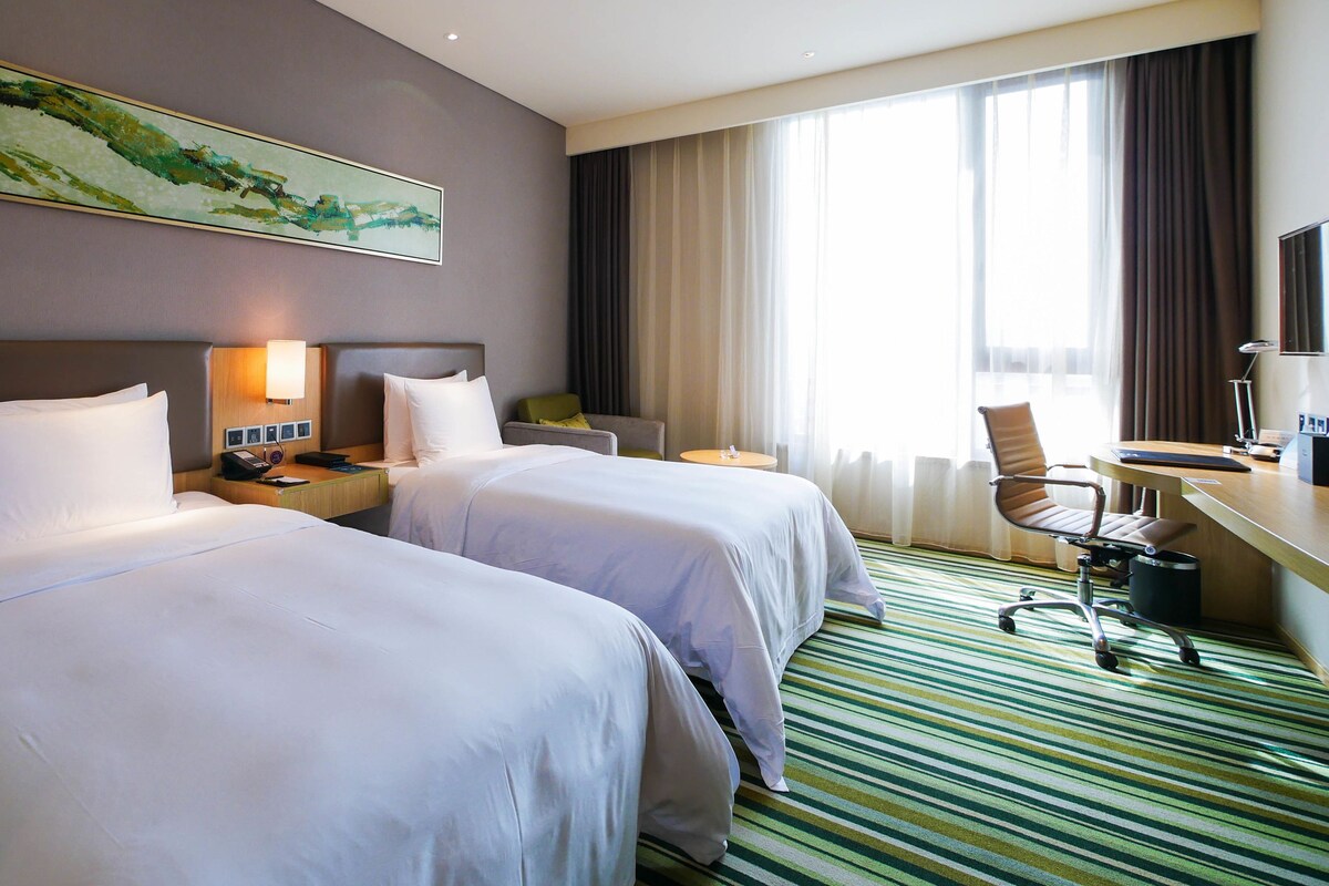 Habitación de hotel con dos camas, un escritorio, una silla, una lámpara y un amplio ventanal con cortinas.