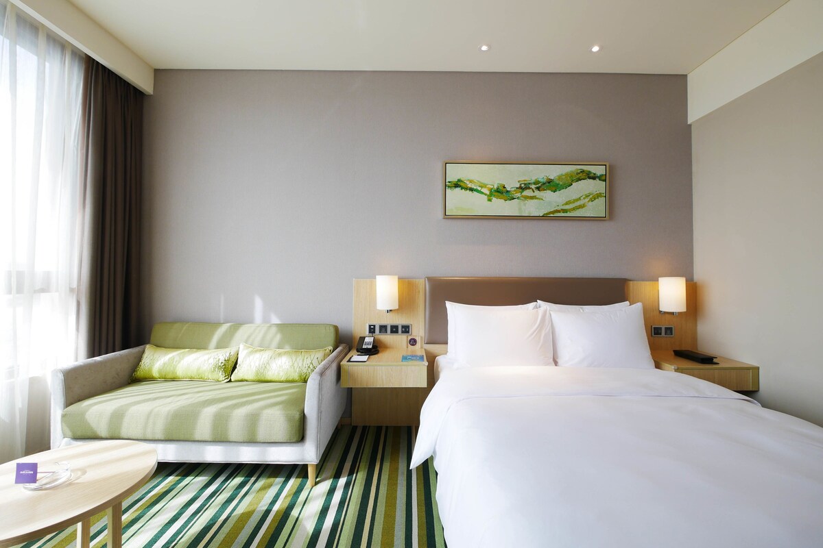 Una habitación de hotel con cama, sofá, mesita de noche, lámpara, teléfono y un cuadro en la pared.