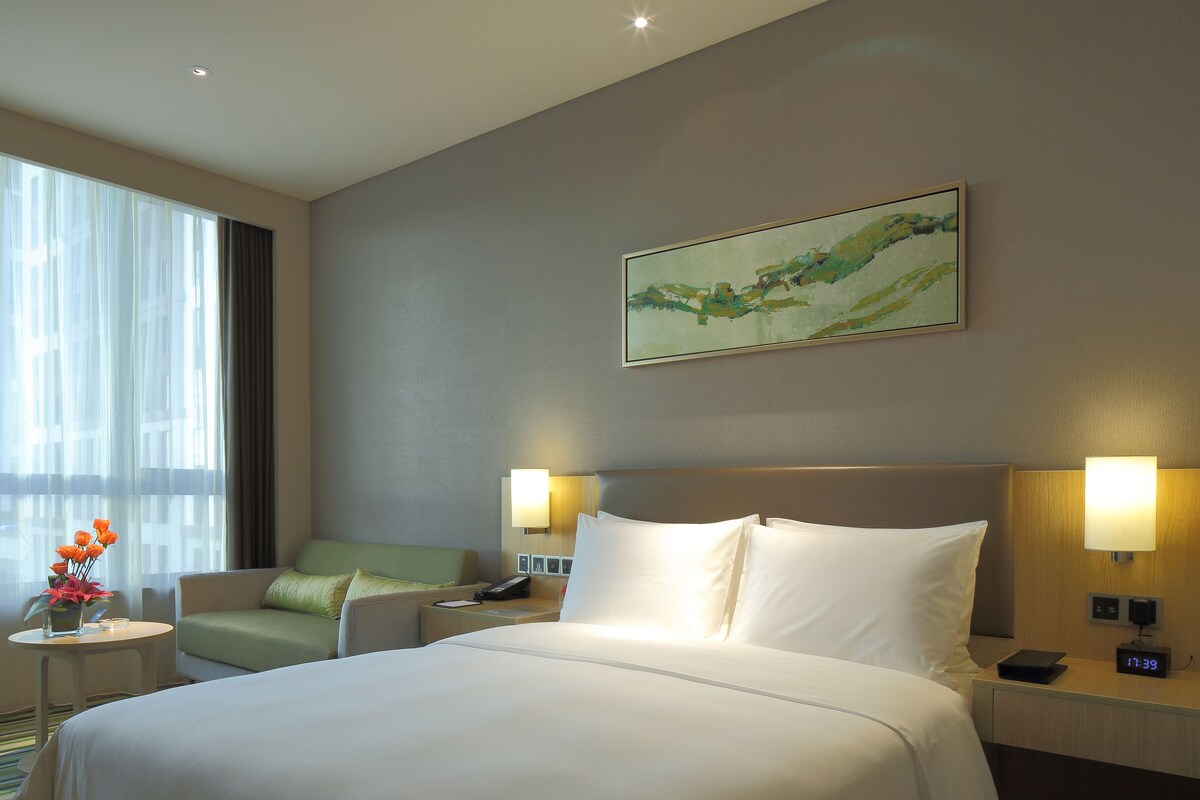 Habitación de hotel con una cama grande, dos lámparas de noche, un cuadro en la pared, una silla, una mesita con un jarrón de flores y una ventana con cortinas.