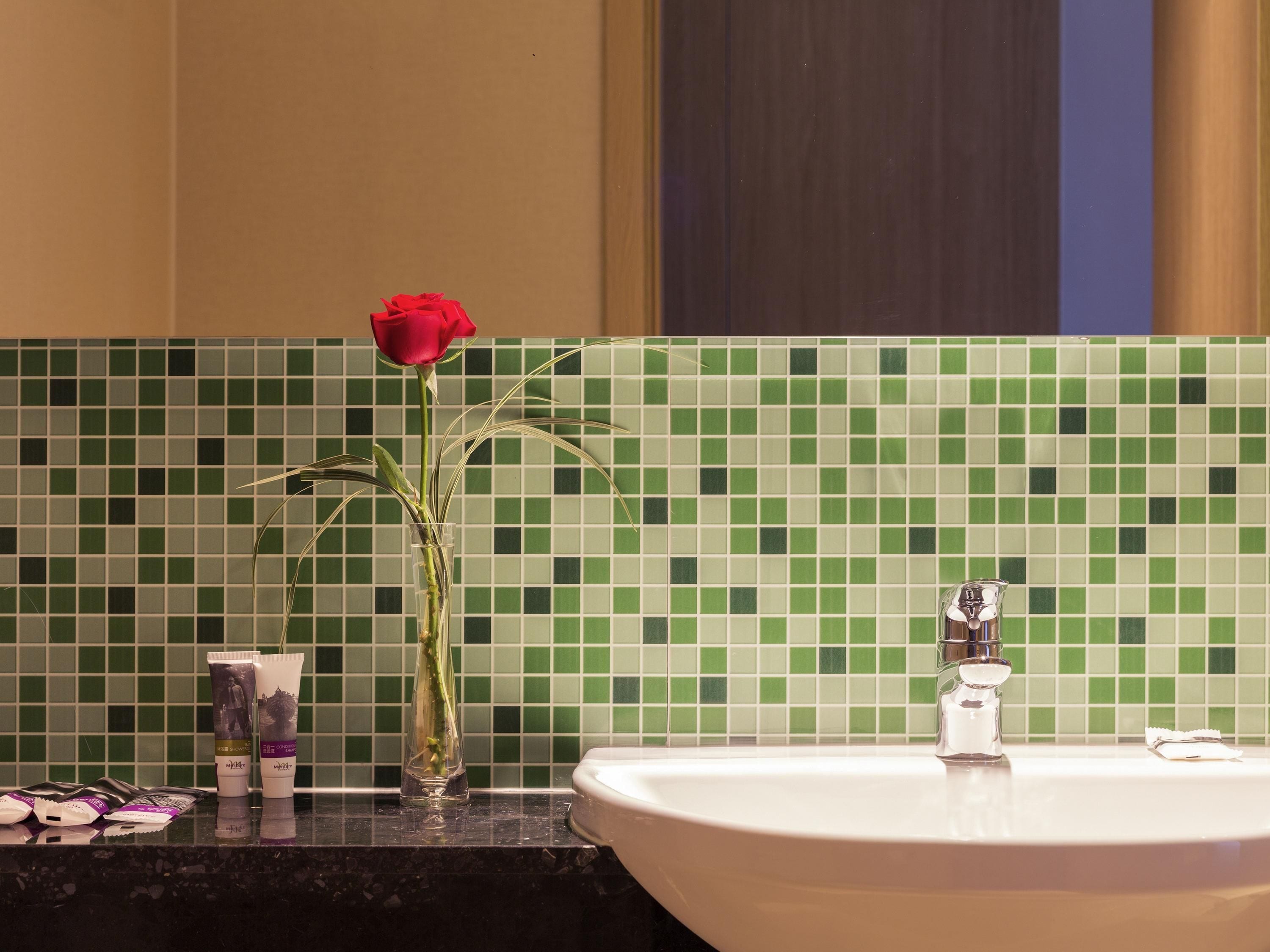 Ein Badezimmer mit Waschbecken, einer Vase mit einer roten Rose und Pflegeprodukten auf der Ablagefläche.