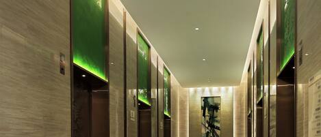 A modern hotel corridor with a green-lit elevator, a patterned carpet, and a minimalist design.