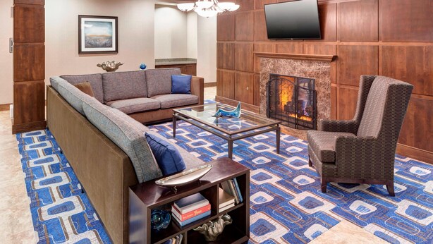 A modern living room with a sofa, armchair, coffee table, bookshelf, fireplace, and a flat-screen TV mounted on the wall. - Residence Inn by Marriott Dallas Plano/Richardson (Plano)
