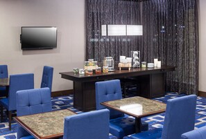 A hotel room with a large flat-screen TV mounted on the wall, a dining area with a long table and blue upholstered chairs, and a patterned carpet. - Residence Inn by Marriott Dallas Plano/Richardson (Plano)