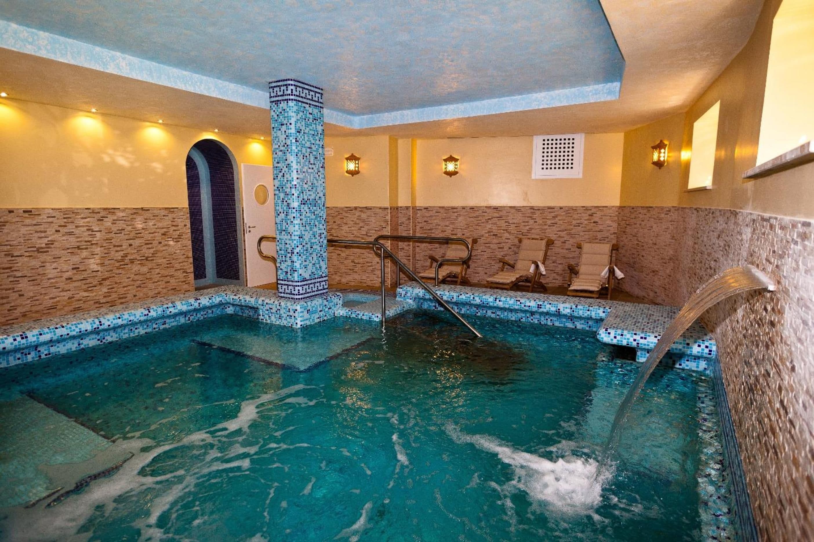 An indoor swimming pool with a mosaic-tiled column, a water fountain, and lounge chairs.