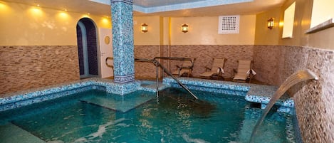 An indoor swimming pool with a mosaic-tiled column, a water fountain, and lounge chairs.