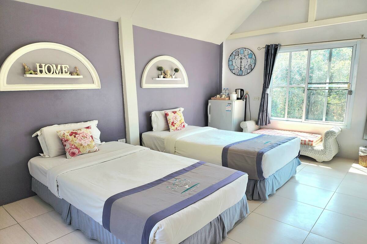 A bedroom with two beds, a window with curtains, a refrigerator, and a wall-mounted clock.