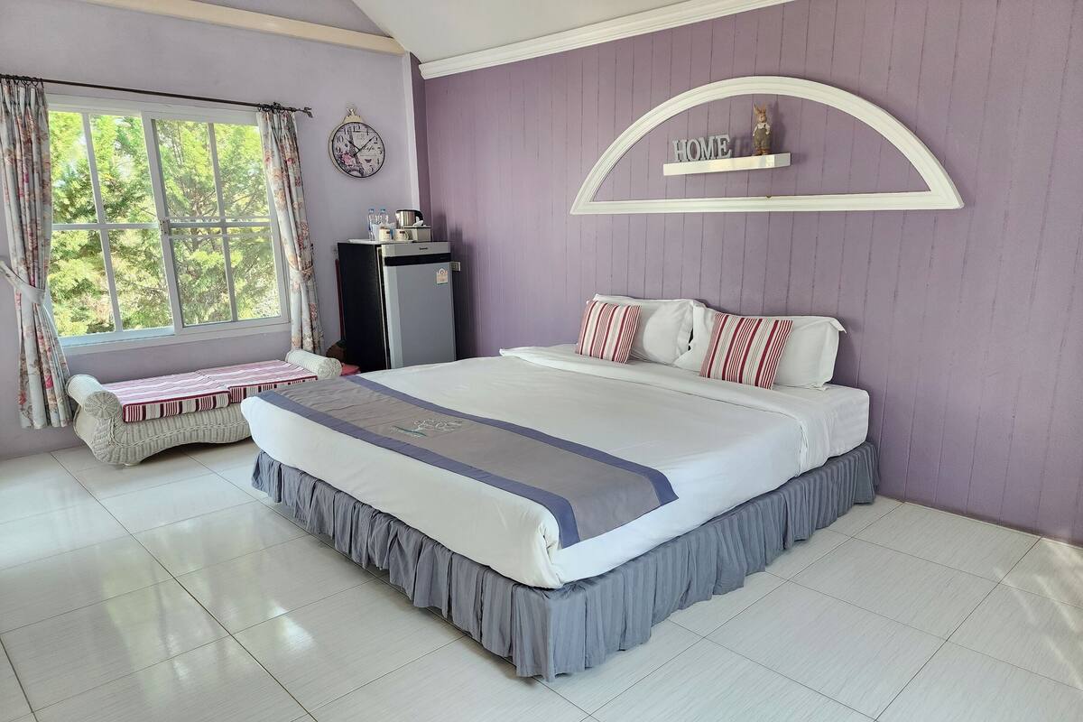 A bedroom with a large bed, a window with curtains, a refrigerator, a clock, and a wall-mounted shelf with a decorative item.
