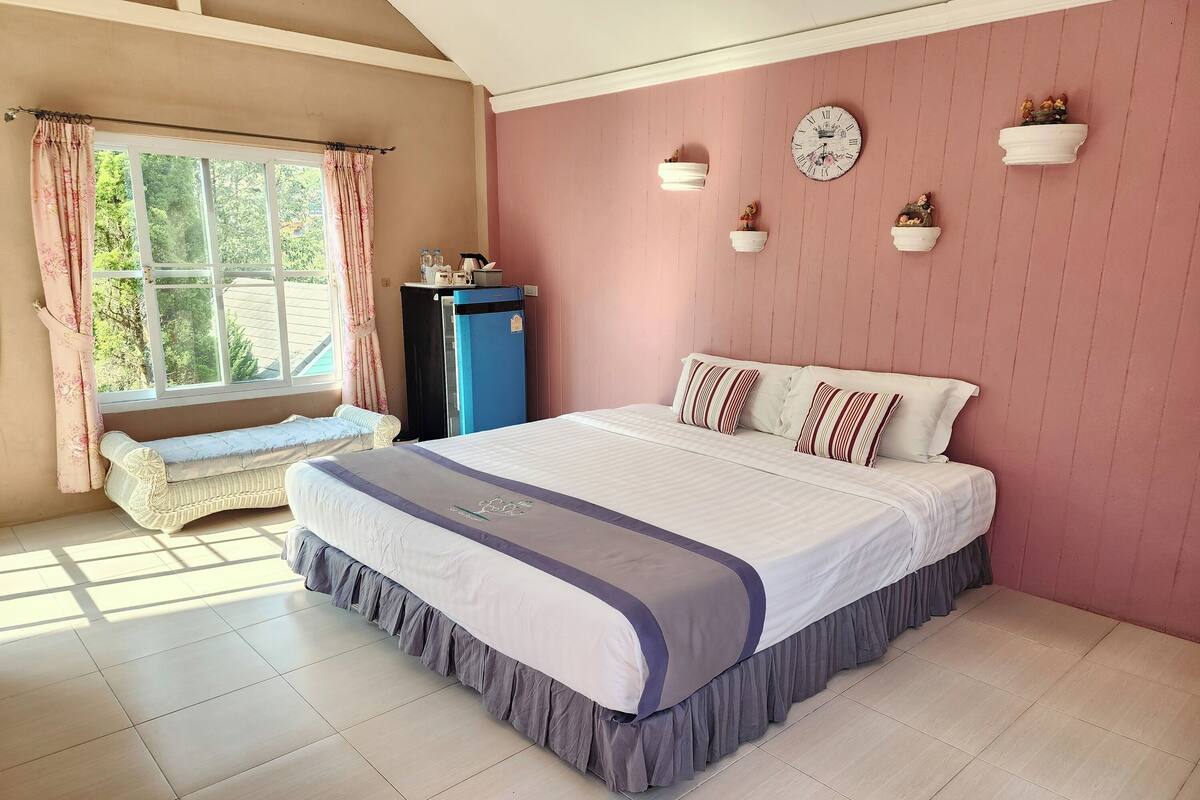 A bedroom with a large bed, a bench, a blue refrigerator, and a window with curtains.