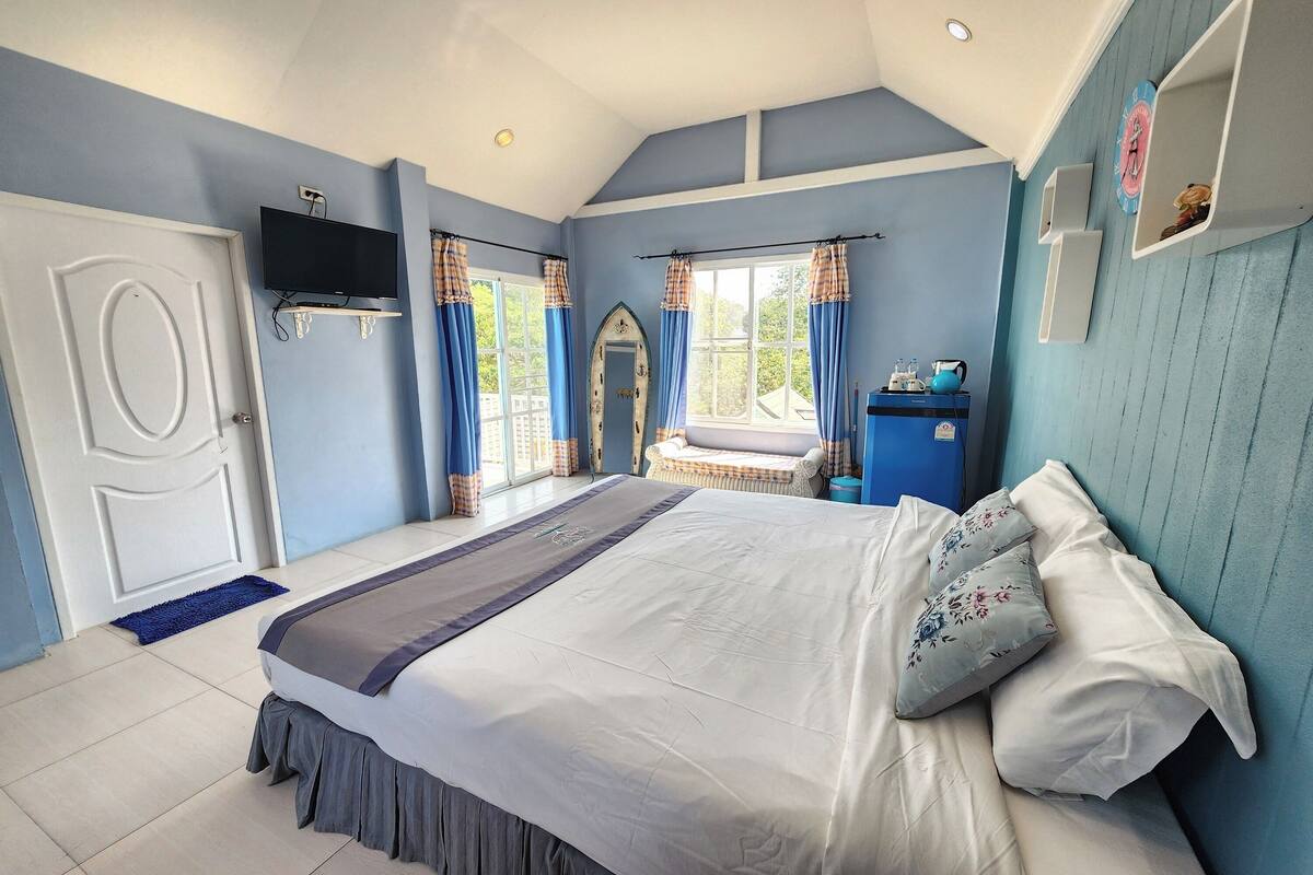 A bedroom with a bed, a TV mounted on the wall, a window with curtains, and a small table with a kettle.
