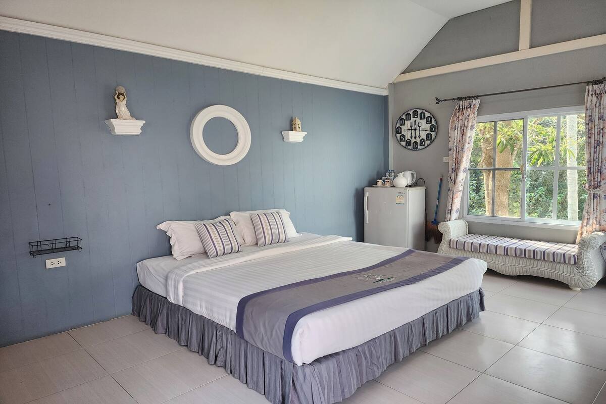 A bedroom with a large bed, a bench, a wall clock, and a window with curtains.