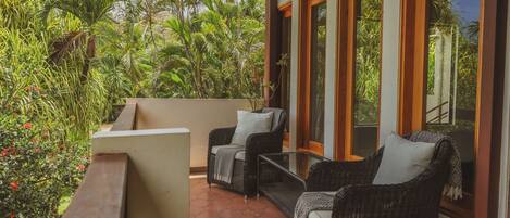A covered outdoor seating area with wicker furniture and large windows overlooking a lush garden.