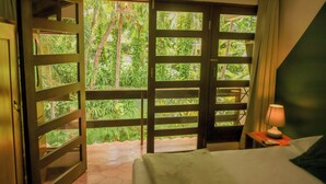 A hotel room with a bed, a nightstand, a lamp, and a view of lush greenery through a sliding door. - Hotel Villa Del Sol (Sardinal)