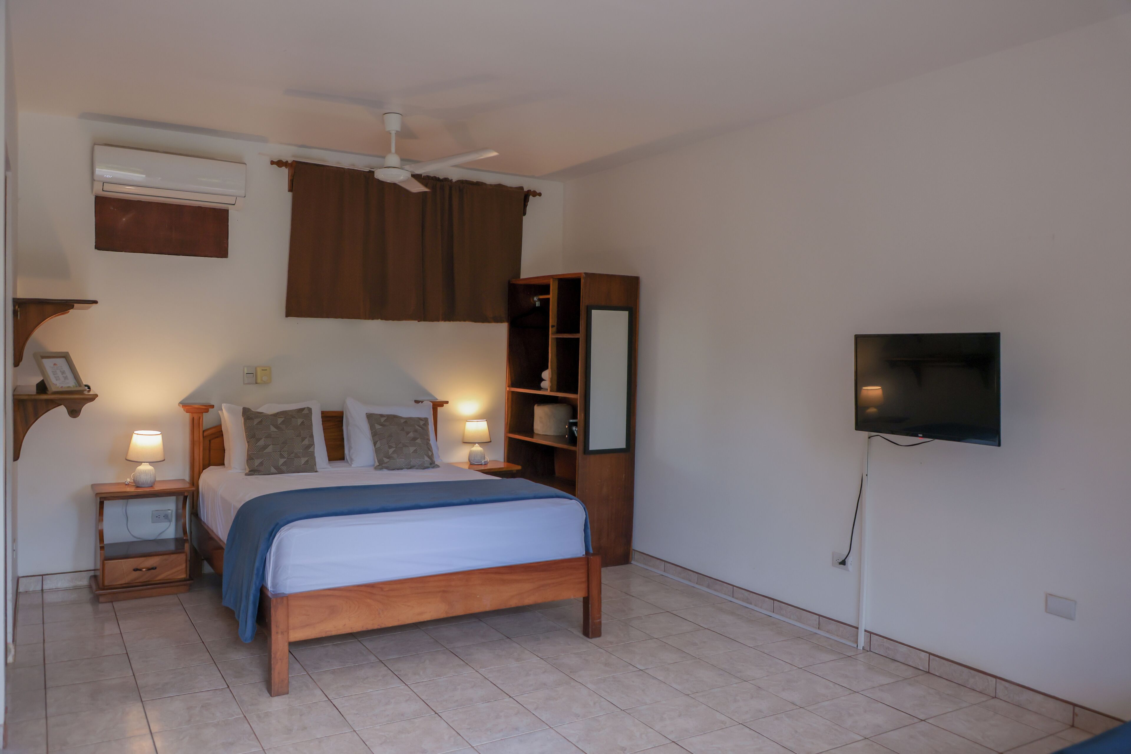 A bedroom with a wooden bed, bedside lamps, a ceiling fan, a TV mounted on the wall, and a brown curtain.