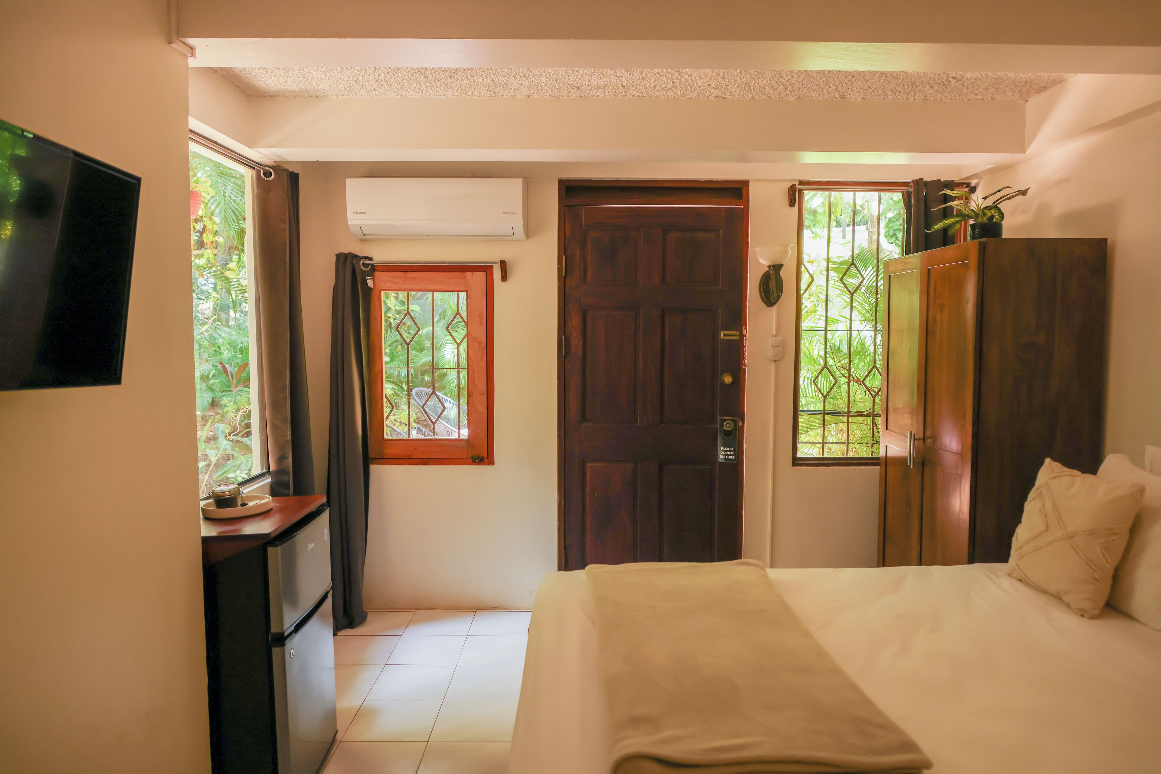 A bedroom with a bed, a TV, a wardrobe, and a window with a view of greenery.