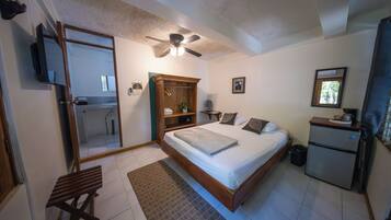 Un dormitorio con cama, ventilador de techo, kitchenette pequeña, banco de madera y baño visible a través de una puerta abierta.