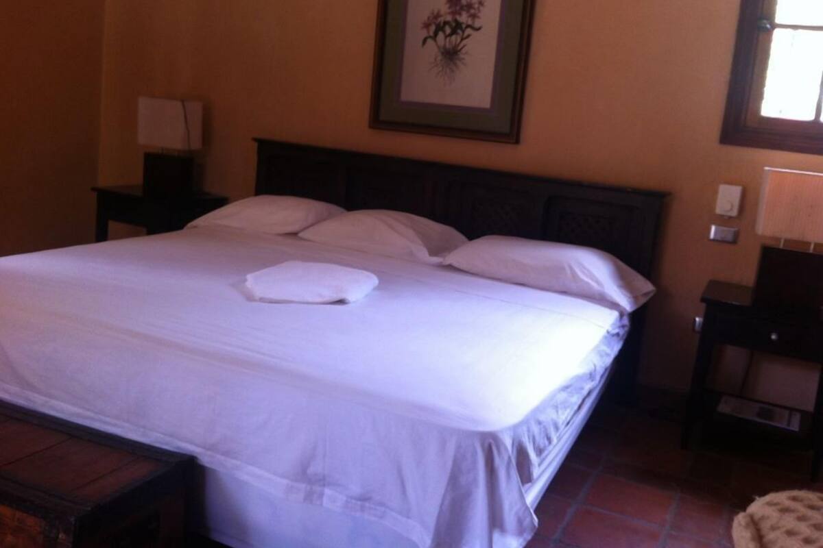A neatly made bed with white linens and a folded towel, a framed floral artwork on the wall, and a nightstand with a lamp.