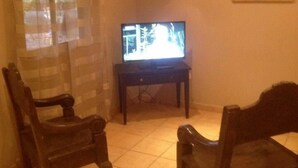 A living room with a television on a stand, two wooden chairs, a small table, and a wall-mounted picture frame. - Cabinas Cigalou Tamarindo (Tamarindo)