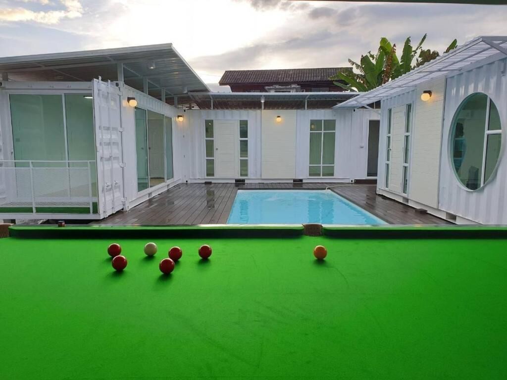 A pool area with a green pool table, a pool, and white walls with large windows.