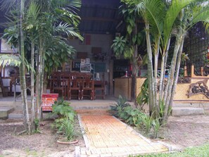 A wooden pathway leading to a building with a signboard, surrounded by palm trees and plants.