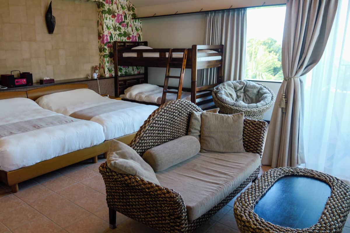 Une chambre d’hôtel avec deux lits, un canapé, un fauteuil et une fenêtre avec des rideaux.