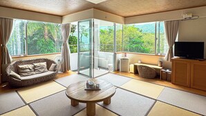 Un salon moderne avec un revêtement de sol en tatami, un canapé, une table avec un plateau, un téléviseur et de grandes fenêtres donnant sur un paysage verdoyant.