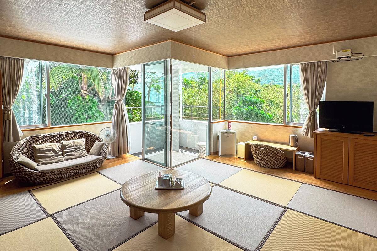 Un salon moderne avec un revêtement de sol en tatami, un canapé, une table avec un plateau, un téléviseur et de grandes fenêtres donnant sur un paysage verdoyant.