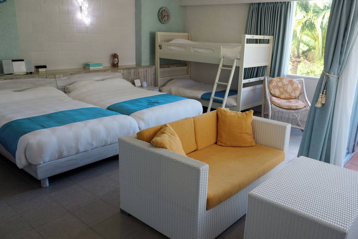 Une chambre d’hôtel avec deux lits, un canapé, une chaise, une échelle et une vue sur la verdure environnante.