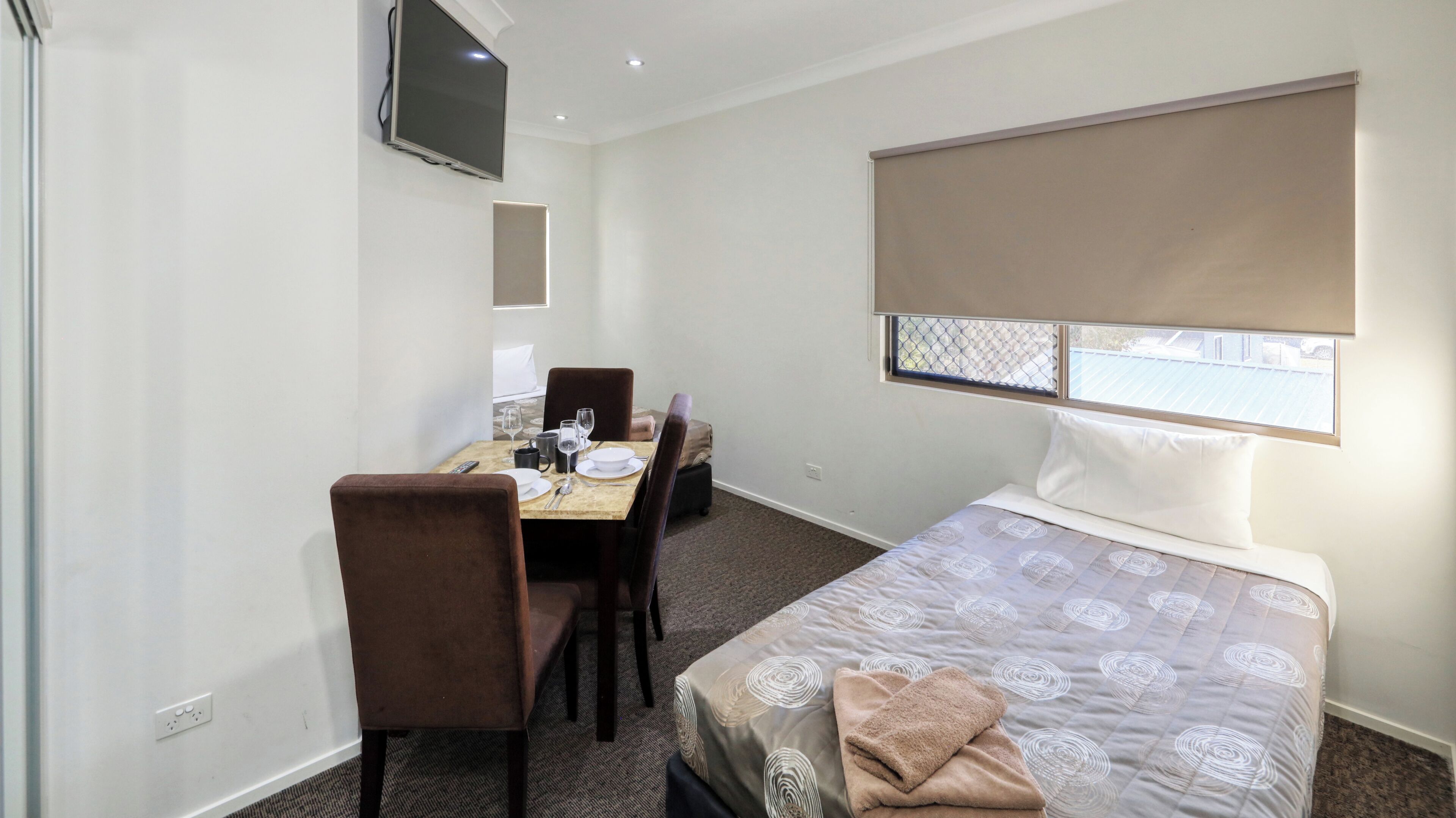 A hotel room with a bed, a dining table with chairs, a TV mounted on the wall, and a window with a view.