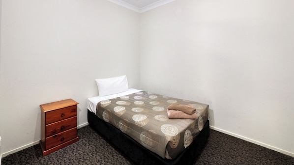 A single bed with a patterned bedspread, a wooden nightstand with drawers, and a plain carpeted floor.