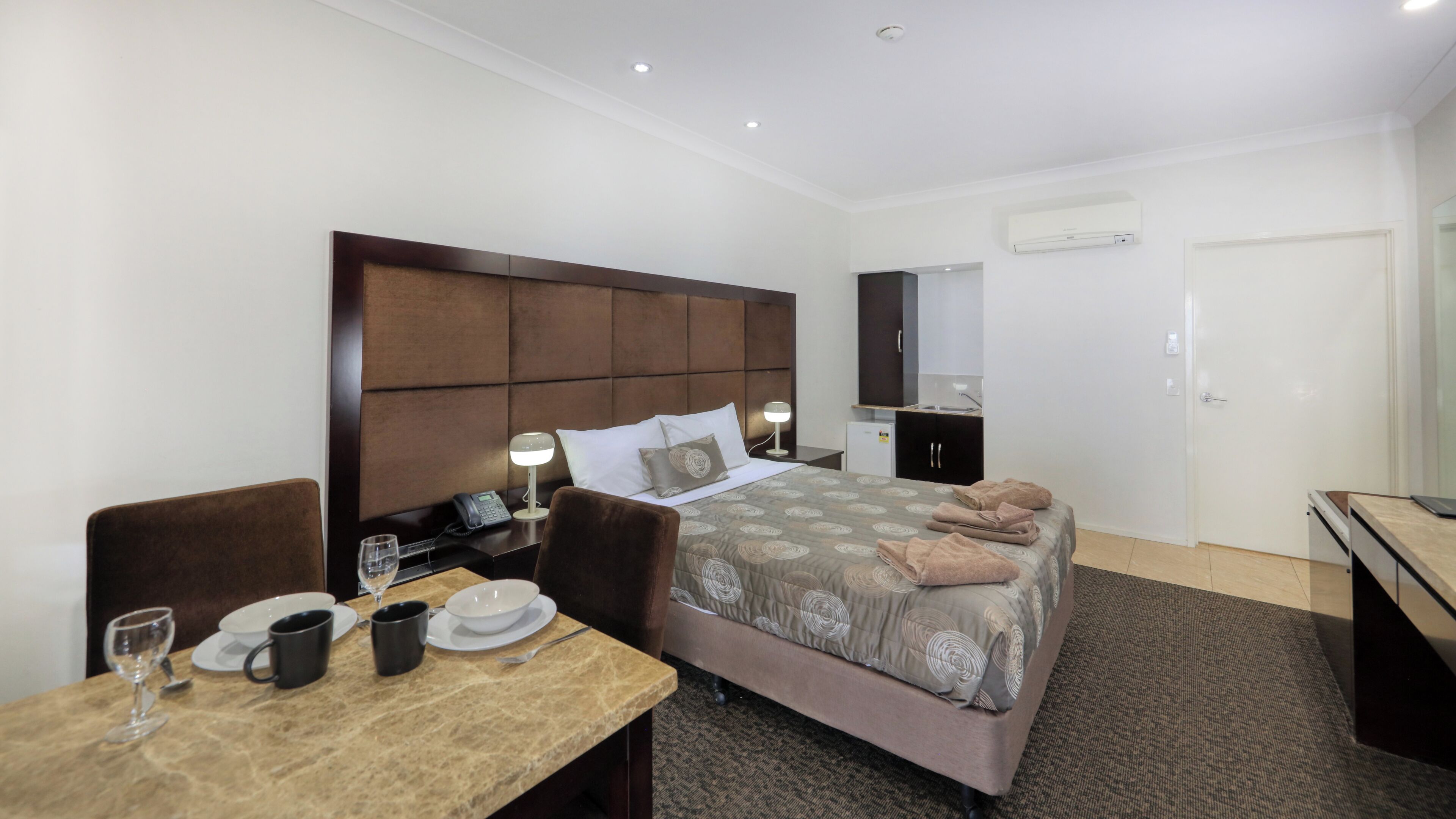 A hotel room with a bed, bedside table, lamp, and a small desk with a chair.