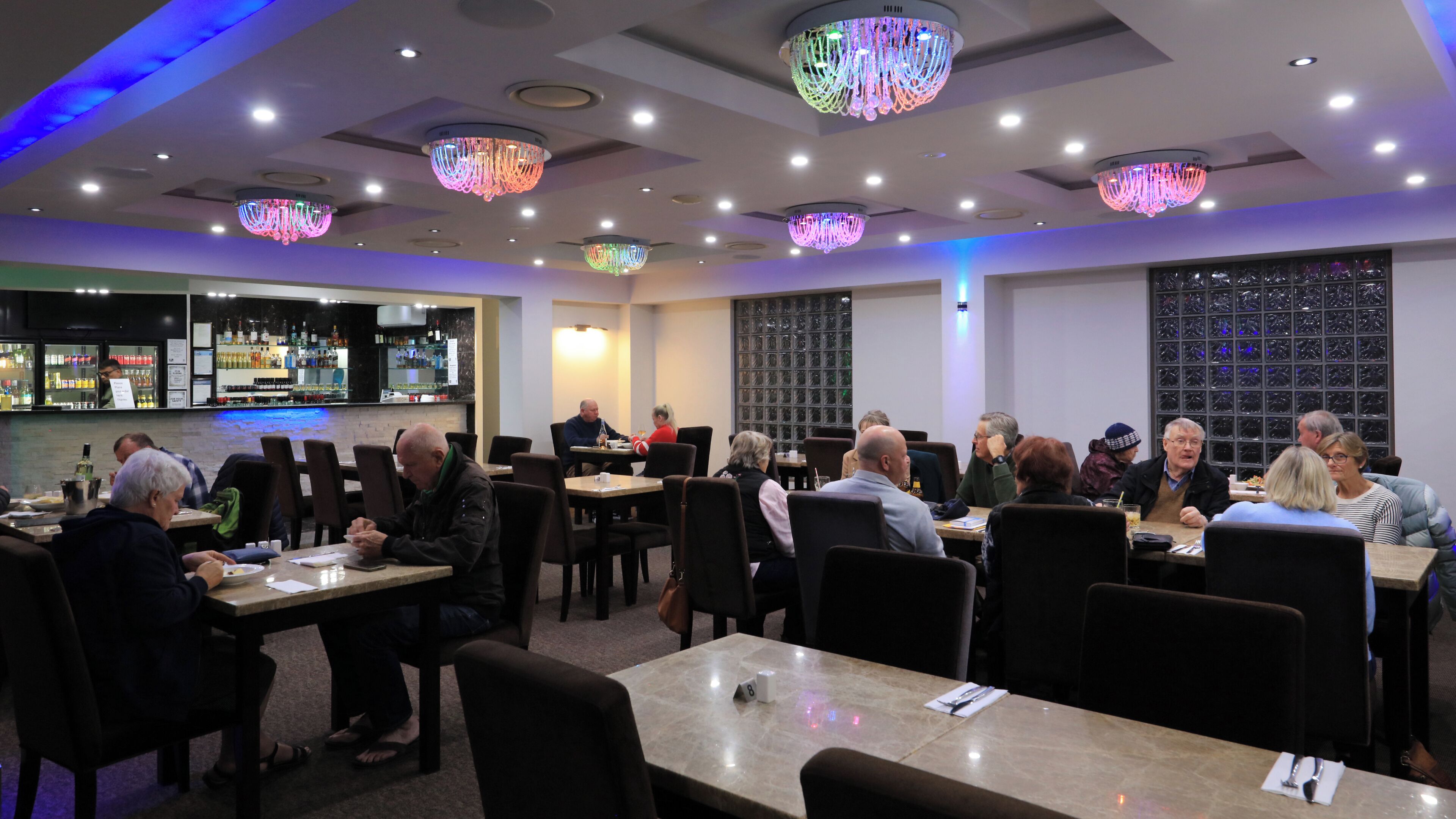 A restaurant with multiple tables and chairs, a bar area with bottles, and decorative chandeliers.
