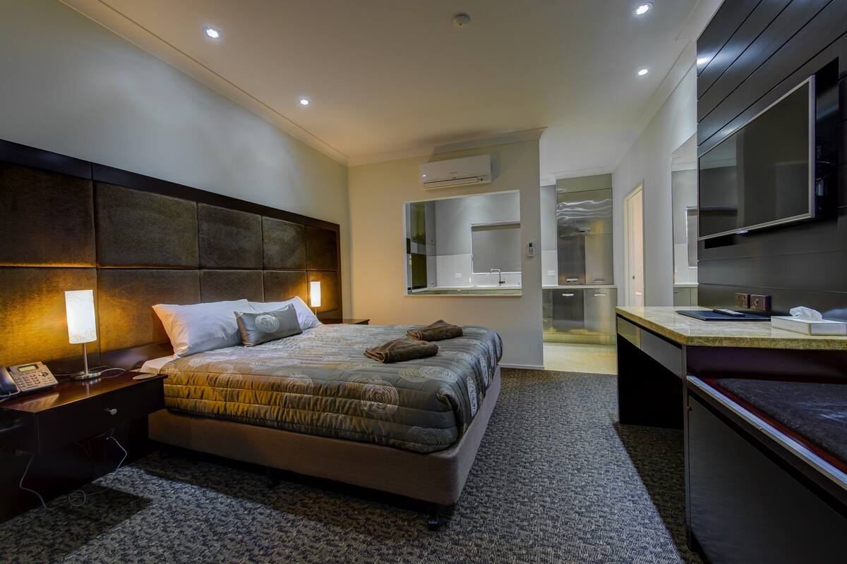 A modern hotel room with a large bed, bedside table, lamp, and a desk with a chair.