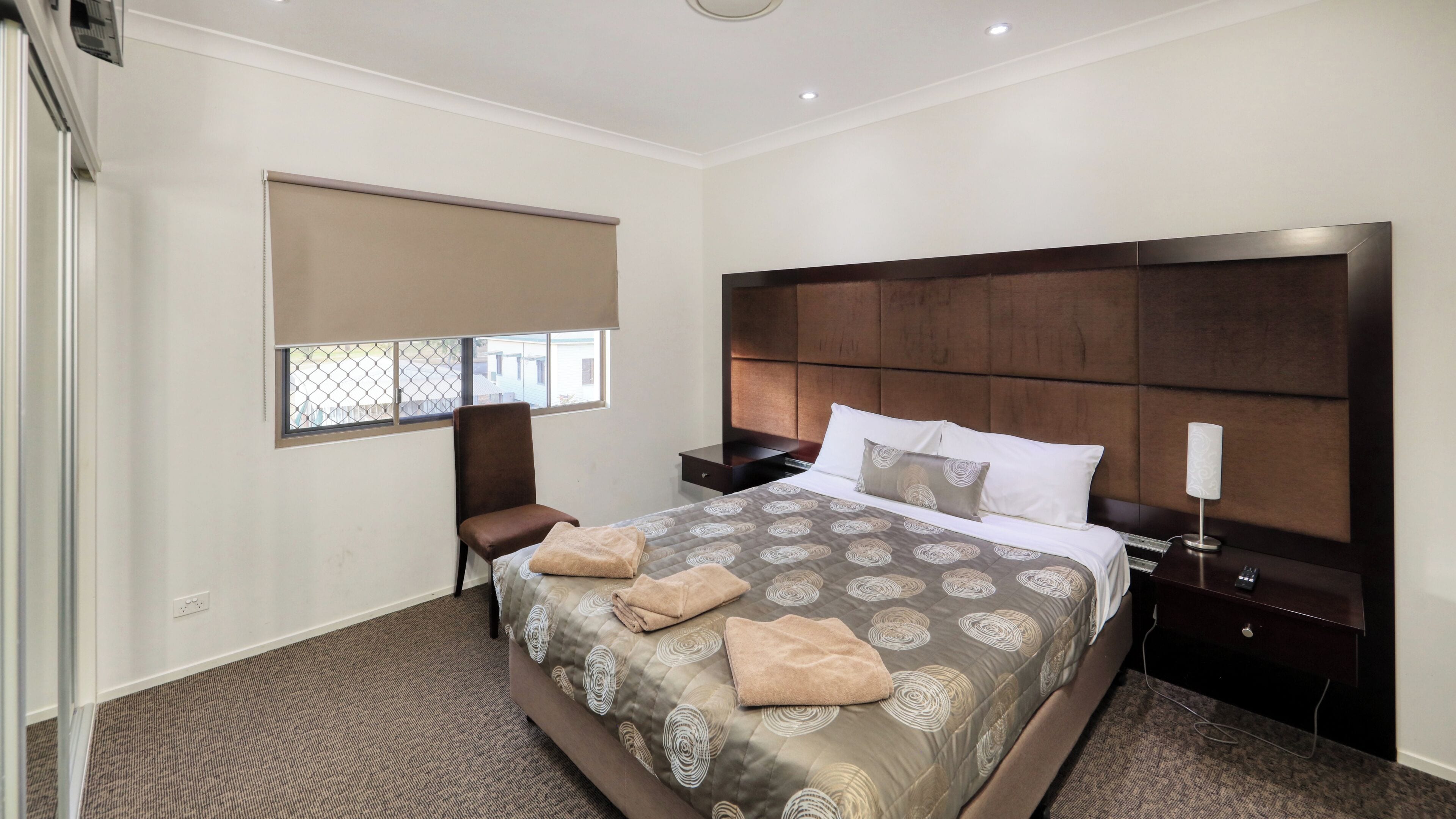 Habitación de hotel con una cama grande, una mesita de noche, una silla, una ventana con persianas y una lámpara fijada a la pared.
