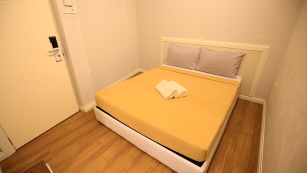 A small, neatly made bed with a yellow mattress and white headboard, a folded white towel on the bed, and a wooden floor. - Travelogue KL - Hostel (Kuala Lumpur)