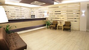 Un área de recepción con un mostrador de madera, dos sillas, una planta en maceta y una pared con listones horizontales.