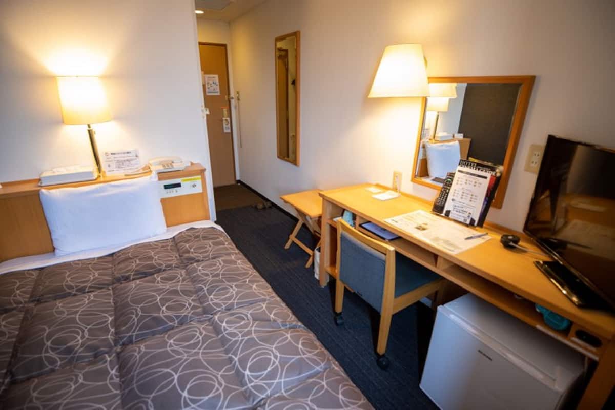 A hotel room with a bed, desk, chair, lamp, mirror, and a small refrigerator.