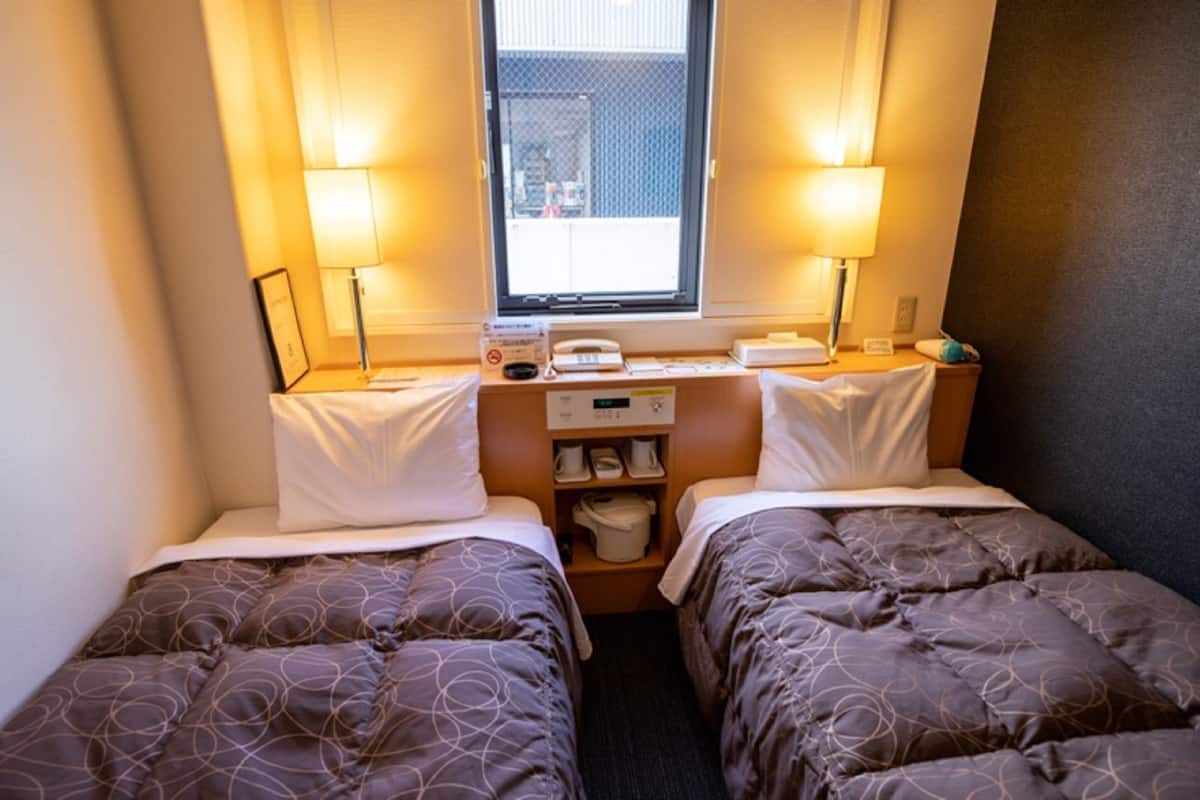 A small hotel room with two beds, a window, a desk with a telephone, and a shelf with toiletries.
