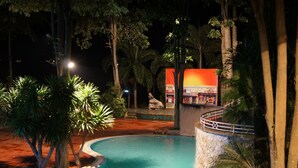 Una zona de piscina nocturna de borde curvo, rodeada de palmeras y un muro de piedra.