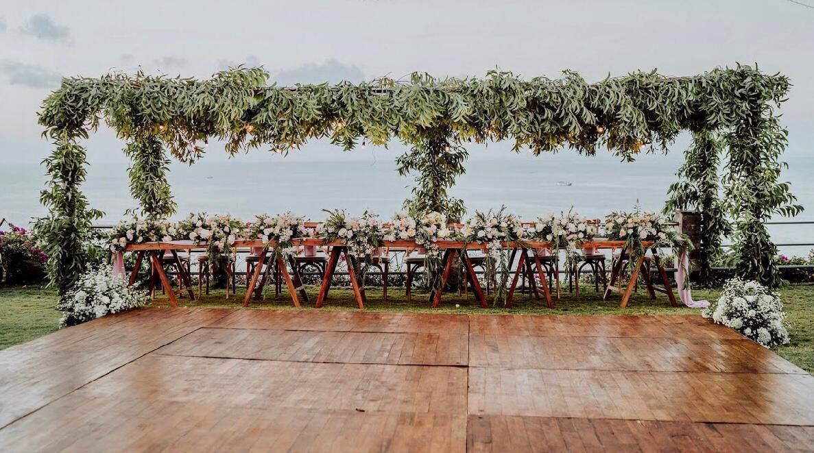Montaje para una boda al aire libre con un escenario de madera, decoración floral y un fondo de follaje.