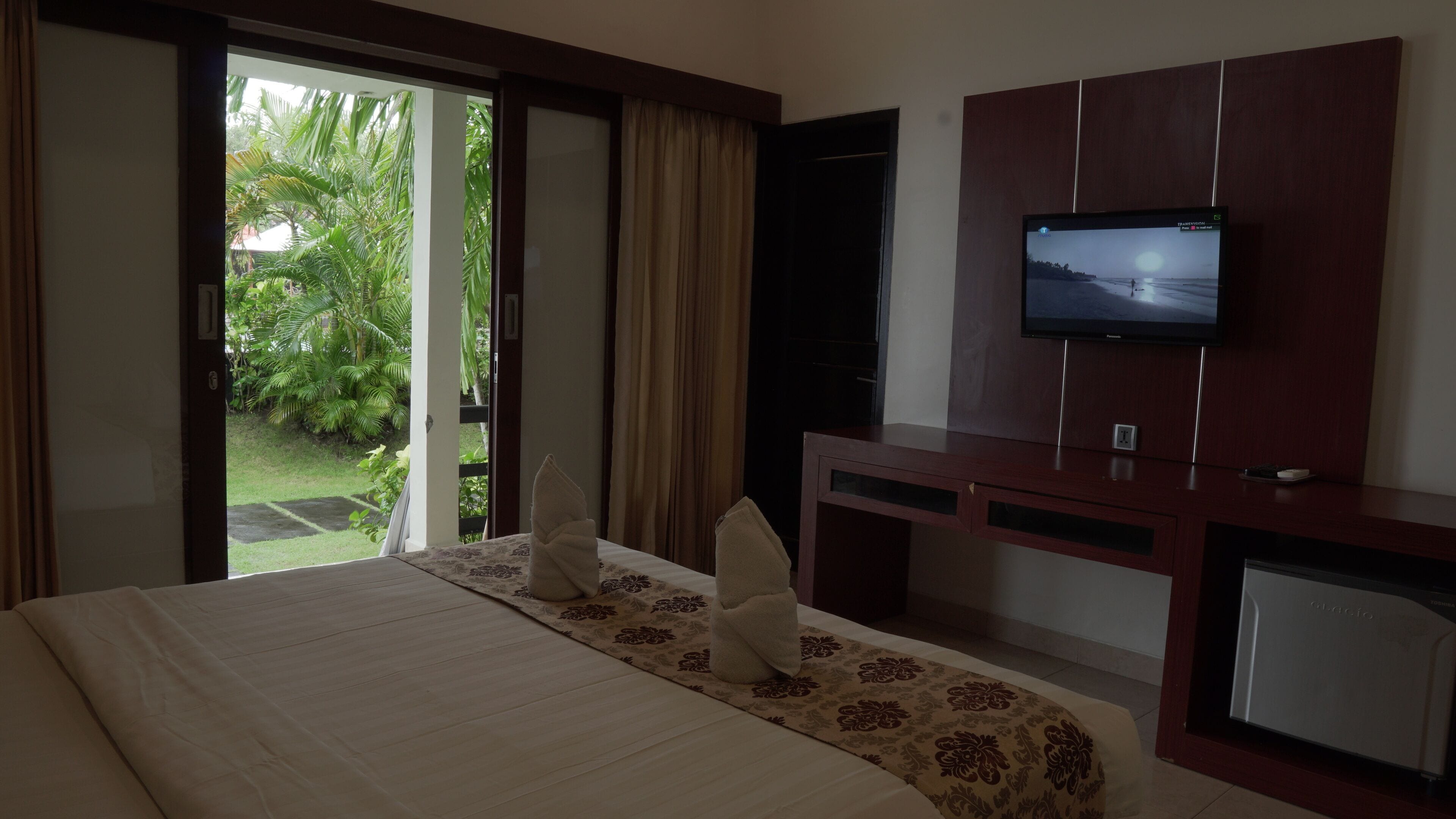 A hotel room with a bed, a TV mounted on a wooden cabinet, and a view of greenery through an open door.