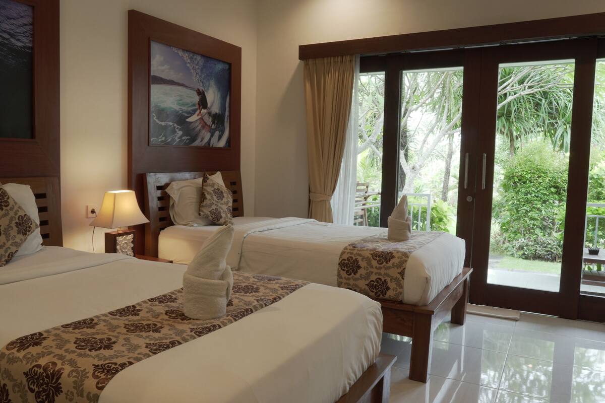 A hotel room with two beds, a painting of a surfer, a window with a view of greenery, and a towel on each bed.