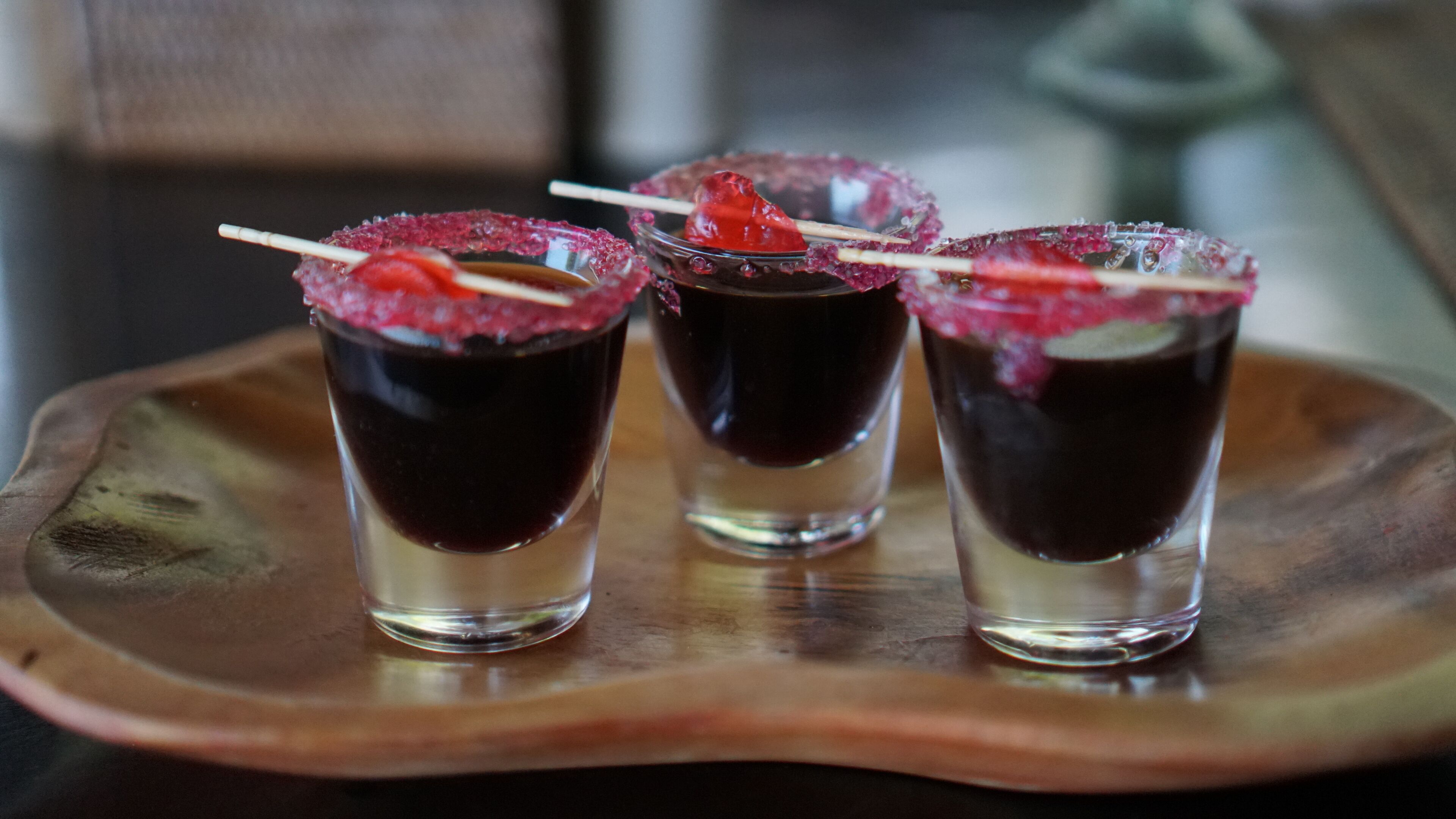 Three shot glasses with dark beverages, garnished with a red fruit and a sugar rim, on a wooden tray.